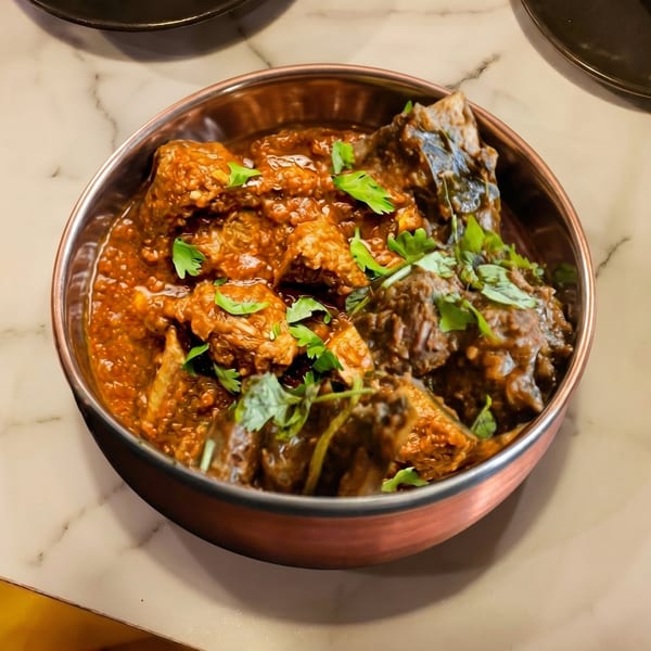A copper bowl filled with rich, dark brown curry, garnished with fresh green cilantro on a marble table.